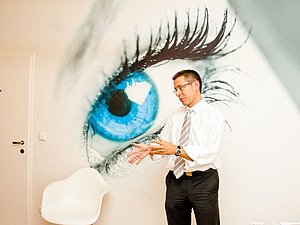 Ein Mann mit Brille steht gestikulierend vor einer Wand auf der ein großes blaues Auge zu sehen ist