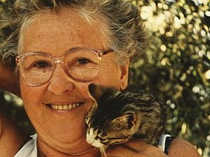 Eine ältere Dame mit Dauerwelle und Brille lächelt fröhlich in die Kamera während sie ein Katzenjunges streichelt, das auf ihrer Schulter sitzt und sich an ihr Gesicht schmiegt