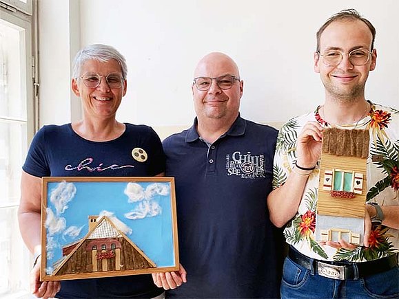 Drei Personen lächeln in die Kamera. Die Frau mit kurzen grauen Haaren links hält eine taktile Fotografie eines Hausdaches vor einem blauen Himmel; die einzelnen Elemente sind aus verschiedenen Materialien wie Watte und Holzstäbchen. Sie trägt eine Brille und einen gelben Button mit drei gelben Punkten. Der Mann in der Mitte trägt eine Brille und hat eine Glatze. Der Mann rechts trägt auch eine Brille und ein bunt gemustertes Hemd, er hält eine taktile Fotografie einer Hausfassade.