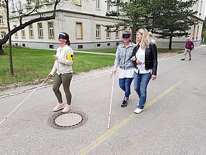 Zwei Frauen sind mit Augenbinde und Langstock auf einer Straße unterwegs. Eine der Frauen wird von einer sehenden Frau begleitet.