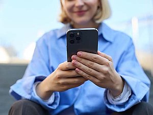Eine blonde Frau mit blauem Hemd sitzt lächelnd da und hält ihr Smartphone vor sich. Hinter ihr sieht man verschwommen den Himmen und Dächer. 