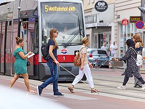 Personen kreuzen an Zebrastreifen die Straße, auch eine junge Frau mit Langstock, sunkler Brille und Rucksack. Dahinter wartet eine Straßenbahn mit der Aufschrift: "5, Westbahnhof"