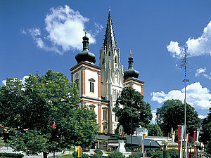 Park mit Bäumen und Blumen, dahinter die Türme der Basilika Mariazell vor blauem Himmel.