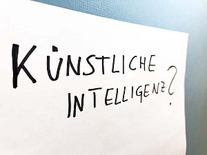 Auf einem weißen Blatt Papier, das an einer blauen Wand hängt, steht in Großbuchstaben: "Künstliche Intelligenz?"