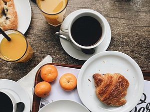 Das Bild zeigt eine Frühstücksszene mit Gebäck, Croissant, Kuchen, Orangensaft, Kaffee und Mandarinen auf einem Holztisch.