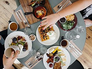 Runder Tisch von oben, auf dem viele Teller mit appetitlichem Frühstück stehen. Verschiedene Hände beugen sich darüber und greifen zu. 