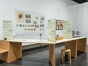 Ein Museumsausstellungsbereich mit einem interaktiven Tisch, der Informationstafeln, eine Hörstation und historische Bilder zeigt. Glasglocken mit Miniaturbäumen und ein Holzschemel ergänzen die Szene.