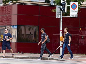 Personen verlassen den Gehsteig um die Straße zu überqueren, eine junge Frau verwendet einen Langstock. 