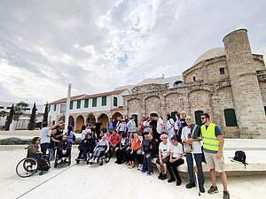 Eine große, diverse Gruppe, darunter Personen mit Seh- und Mobilitätseinschränkungen, posiert für ein Gruppenfoto vor einem historischen Steingebäude mit Rundbögen und Kuppeln. Der Himmel ist bewölkt, einige Teilnehmer sitzen in Rollstühlen, andere stehen oder gestikulieren.