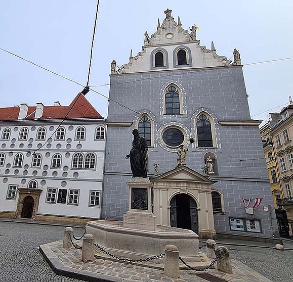 Blaue Kirche im Renaissancestil mit gotischen Elementen, davor ein Brunnen mit Statue in der Mitte. 