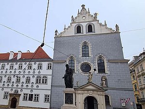 Blaue Kirche im Renaissancestil mit gotischen Elementen, davor ein Brunnen mit Statue in der Mitte. 