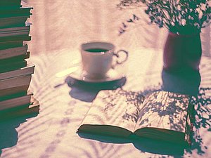 Bücherstapel, Kaffeetasse und aufgeschlagenes Buch stehen auf einem Tisch, Blumen in einer Vase werfen einen Schatten auf das Buch. 